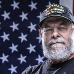 Close-up headshot of an authentic 67 year old United States Navy Vietnam War military veteran looking at the camera. Plenty of copy space on the USA flag stars and stripes red, white and blue background. He is wearing an inexpensive, non-branded, generic, souvenir shop replica Vietnam veteran commemorative baseball hat style cap.